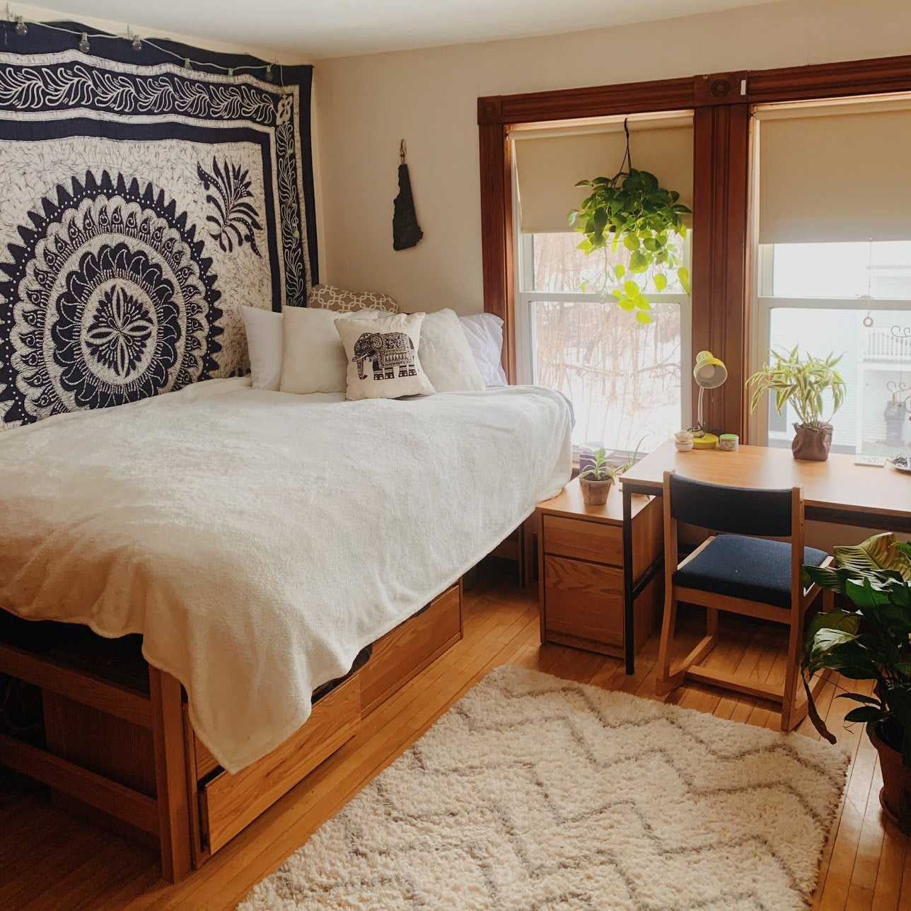 Cozy college dorm room with a Cornell Bigger Bed full-size upgrade, white bedding, wooden furniture, tapestry wall art, desk with plants, and natural light from large windows