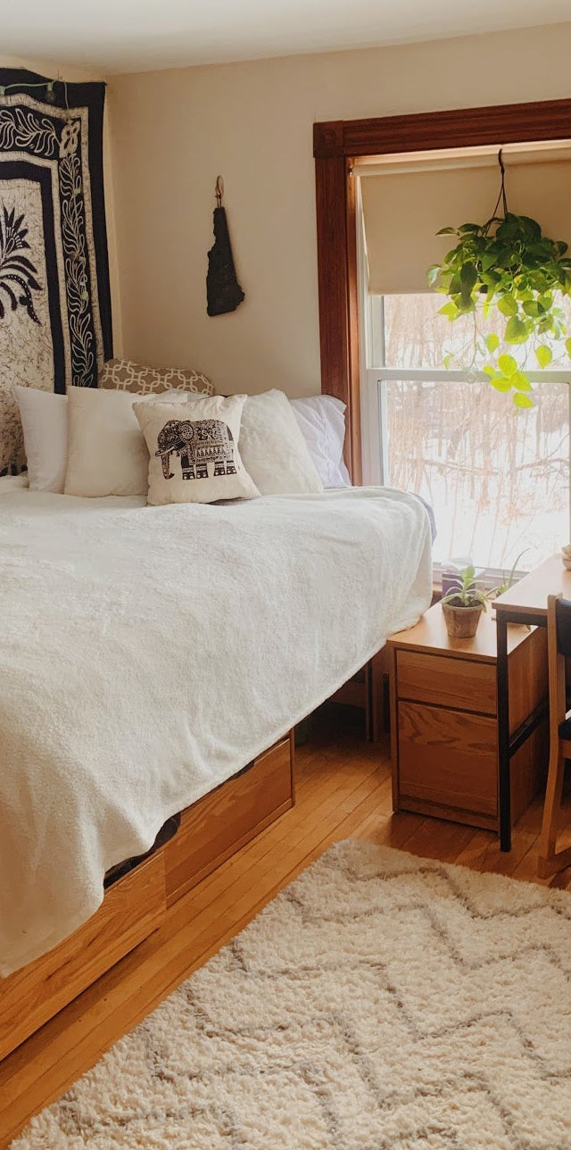 Cozy college dorm room with a Cornell Bigger Bed full-size upgrade, white bedding, wooden furniture, tapestry wall art, desk with plants, and natural light from large windows